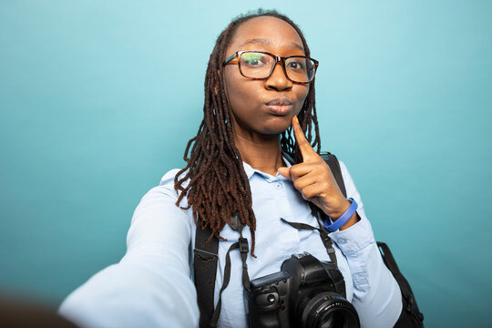 Young african american woman stands confidently with her dslr camera, hand on chin, lost in thoughts. Black female photographer wears a backpack and blue shirt on isolated blue background.