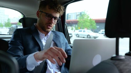 Tired young man in suit finishing work on laptop while sitting in back seat taxi car, working online. Business, trip, transport and technology concept. Slow motion © Vlad