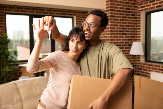 Diverse couple presenting house keys and carrying cardboard box during relocation. Boyfriend and girlfriend celebrate becoming homeowners and new beginnings in modern apartment on moving day.