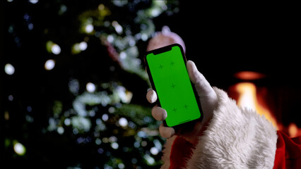 Santa Claus hand in a white glove holds a smartphone with a green screen chroma key background in front of a fireplace and a Christmas tree. A warm festive atmosphere for digital or themed mockups.