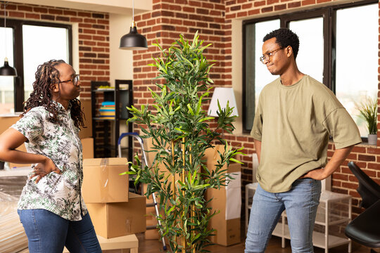 African american couple standing with hands on hips, exchanging looks while planning where to place houseplant in new home. Black woman and man surrounded by boxes, preparing their living space.
