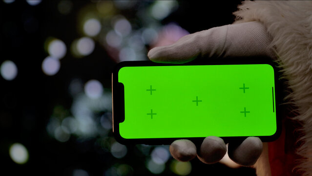 Santa Claus hand in a white glove holds a smartphone with a green screen chroma key background in front of a fireplace and a Christmas tree. A warm festive atmosphere for digital or themed mockups.