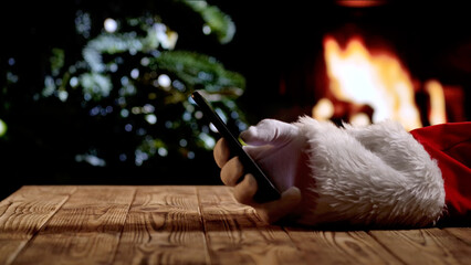 Santa Claus in a red suit and white gloves looks at information on smartphone against the backdrop of fireplace and a Christmas tree with lights. A warm festive atmosphere for New Year or Christmas.