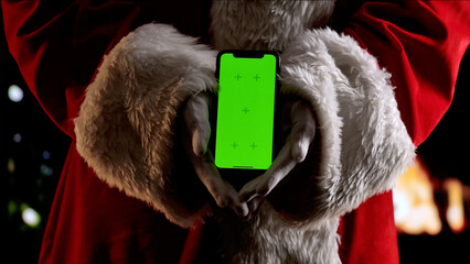 Santa Claus hands in a red suit and white gloves hold a smartphone with a green screen chroma key. A warm festive atmosphere for digital or themed mockups.