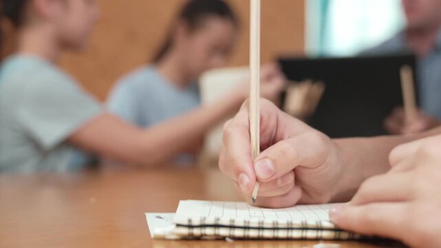 Closeup of student hand taking a note while girl ask teacher a question. Diverse student learning and study with smart happy instructor in classroom. Focus on hand. Blurring background. Edification