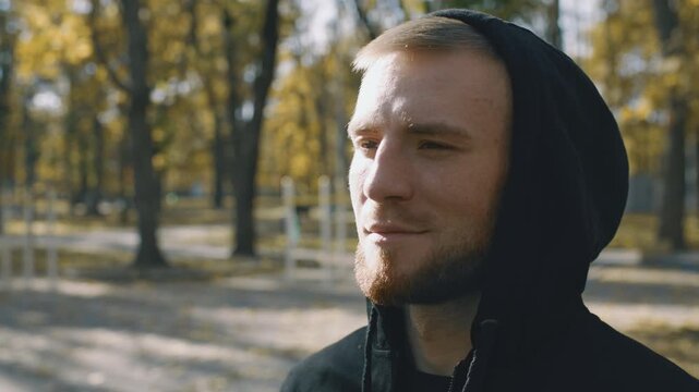 A young man with a beard wearing a hoodie is jogging outdoors. Surrounded by trees, he embraces a healthy lifestyle in a vibrant park. His focus reflects dedication to fitness and well-being.