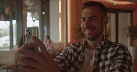 A young business professional uses his smartphone for selfies and digital interaction in a contemporary cafe lounge. This video captures modern work habits and casual social media engagement. - Powered by Adobe