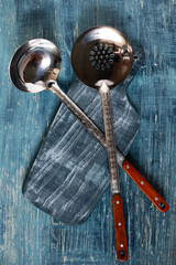 Overhead view of two metal ladles with red handles on a rustic blue wooden background. The utensils...