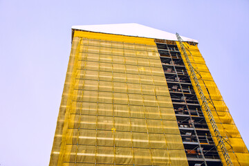 Towering building under construction stands tall, draped in yellow scaffolding netting against blue sky. Its peaked white roof contrasts sharply with the exposed metal frameworks and ladders below