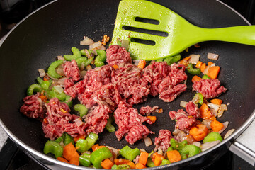 A skillet frying pan of ground beef with celery and carrot pieces with a spatula stirring