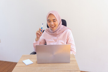 Happy young indonesian hijab woman with pen working on laptop