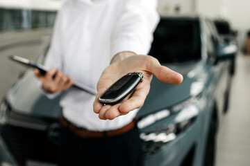 Professional man in white shirt presenting car keys with smartphone in hand, standing in modern car dealership, showcasing vehicle sales and customer service experience