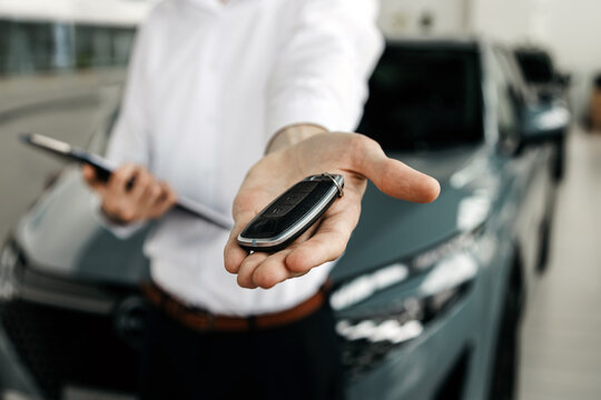 Professional man in white shirt presenting car keys with smartphone in hand, standing in modern car dealership, showcasing vehicle sales and customer service experience