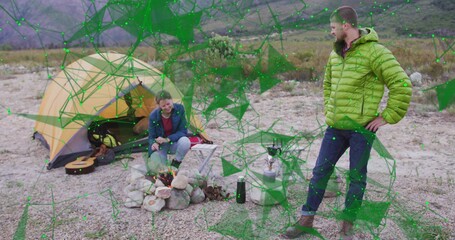 Crouching woman in blue jacket tending firepit while partner observing at campsite with guitar case