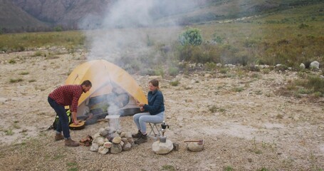 Couple tending campfire meal in rocky clearing, with yellow dome tent, cooking pot and guitar