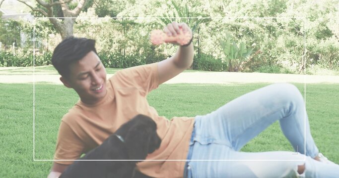 Reclining Asian man wearing tan shirt holding pink bone-shaped toy above black puppy in garden - Powered by Adobe