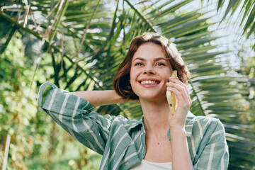 A smiling woman talks on a mobile phone outdoors, framed by lush greenery and bright sunlight.