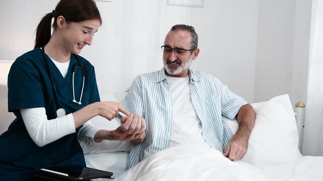 young nurse in uniform takes care of an elderly man, holding his hand and asking him about his condition and giving him encouragement. A nurse uses a stethoscope to check his pulse in a nursing home.
