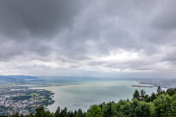 Wide panoramic view from Pfänder mountain showing Lake Constance, coastal towns, and the surrounding landscape under dramatic clouds. 