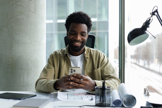 Young joyful black male architect using smartphone at desk in office
