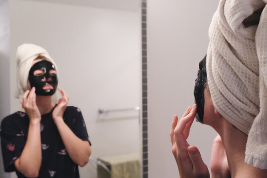 A woman applying a black face mask while looking in the mirror — a calm moment of skincare and self-care captured in a soft, natural light.