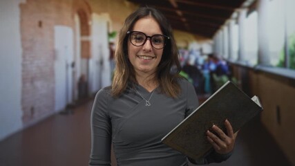 Woman holding book with right hand and wearing glasses in building terrace corridor; thoughtful study.