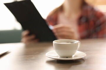Coffee break. Cup of aromatic drink on wooden table and woman with clipboard indoors, selective focus
