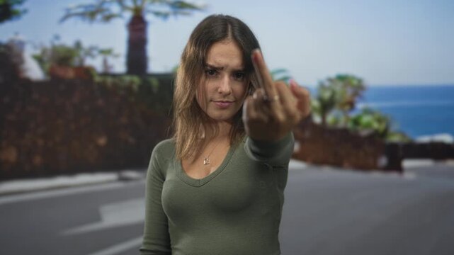 Young hispanic woman showing middle finger and pointing index finger toward camera on street by seaside wall; bold defiance.