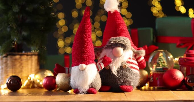 Christmas gnomes, gift boxes and festive decor on wooden table against dark background with blurred lights, changing direction of light. Bokeh effect