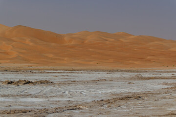 The harsh, cracked white salt flat (Sabkha) with the slopes of the sand dunes