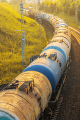 Railway transport of petroleum products travels through green hills at sunset, delivering gasoline and diesel fuel. A freight train with oil tanks rides at sunset in nature.