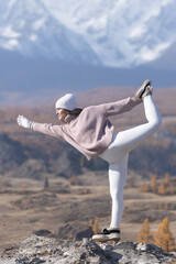 A woman balances elegantly on one leg on a rock in the mountains, showcasing strength and focus. The colorful autumn landscape contrasts beautifully with the snowy peak backdrop.