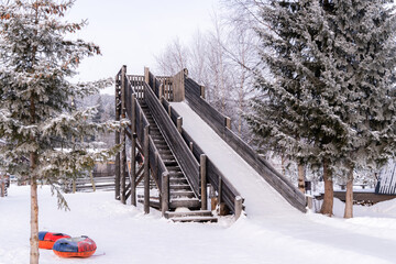 A large wooden slide stands prominently at a snowy playground, flanked by tall pine trees. Colorful inflatable tubes rest on the ground, inviting children to enjoy sledding down the slide.
