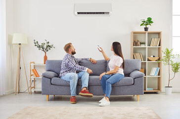 Young family couple sitting on sofa in living room and turning on air conditioner. Happy man and...