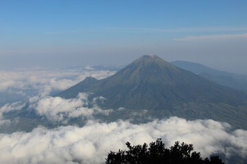 Majestic view of a volcanic mountain peak emerging above a sea of clouds, with green vegetation covering its slopes and a slightly barren summit hinting at geological activity