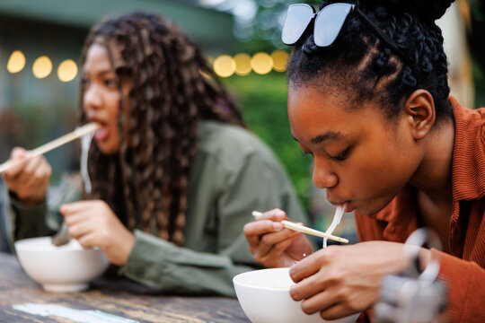 Two women enjoy noodles outdoors with string lights.