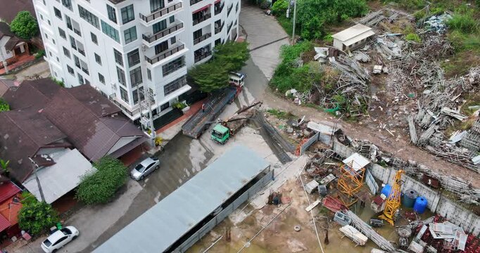 Seen from above, long steel beams are being unloaded from a flatbed by workers using a crane truck in a yard located right beside a modern apartment building.
