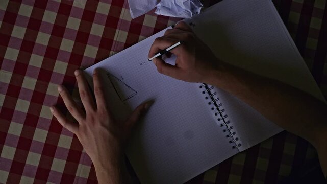 A dedicated student's hand writes a complex math formula and sketches a geometry triangle in a checkered notebook. This close-up captures the intense focus and hard work of an academic study session.