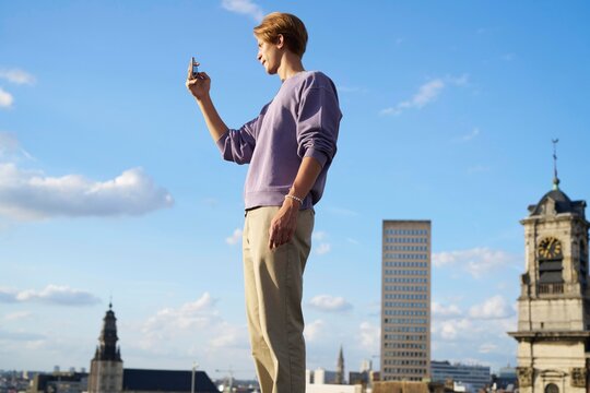 Man Taking Photo on Rooftop With Brussels Skyline Under Blue Sky