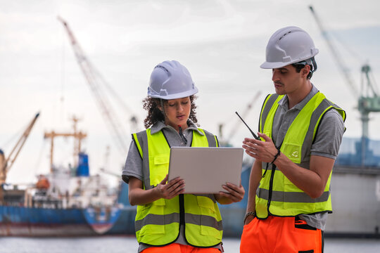 Two engineers wearing safety vests and helmets inspect port operations near the waterfront. They use a laptop and walkie-talkie, symbolizing teamwork, logistics, and marine industry work.
