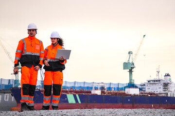 Two port engineers in high-visibility orange uniforms and hard hats communicate while inspecting...