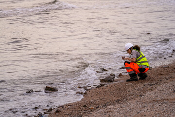 Female environmental scientist squats near shoreline, using smartphone and tablet to record water pollution data, highlighting environmental monitoring and coastal research.