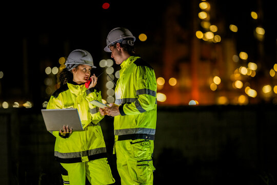 A diverse team of engineers, a man and a woman, in hard hats and safety vests. They are collaborating on a laptop and tablet during a night shift at an industrial site.