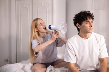 Fototapeta premium A white woman screams into a megaphone at her husband who is sitting on the bed and not paying attention, while the wife screams into a loudspeaker to make him listen. Family conflict.