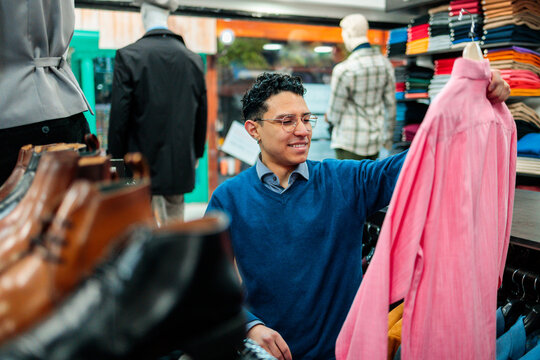 Smiling man holding pink shirt in menswear store - Powered by Adobe