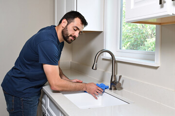 Obraz premium Man doing household chores, diligently scrubbing a white kitchen sink and chrome faucet with a blue sponge for a deep clean