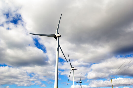 Windfarm with huge mills beside the power poles with electricity lines in the background of blue sky with white clouds.