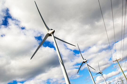 Windfarm with huge mills beside the power poles with electricity lines in the background of blue sky with white clouds.