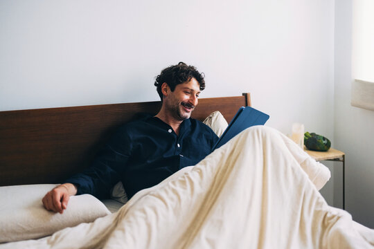Man Relaxing in Bed Reading on a Digital Tablet with a Smile