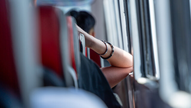 A close-up of a passenger's arm, wearing a beaded bracelet, resting on the back of a red bus seat while they look thoughtfully out the window during travel.